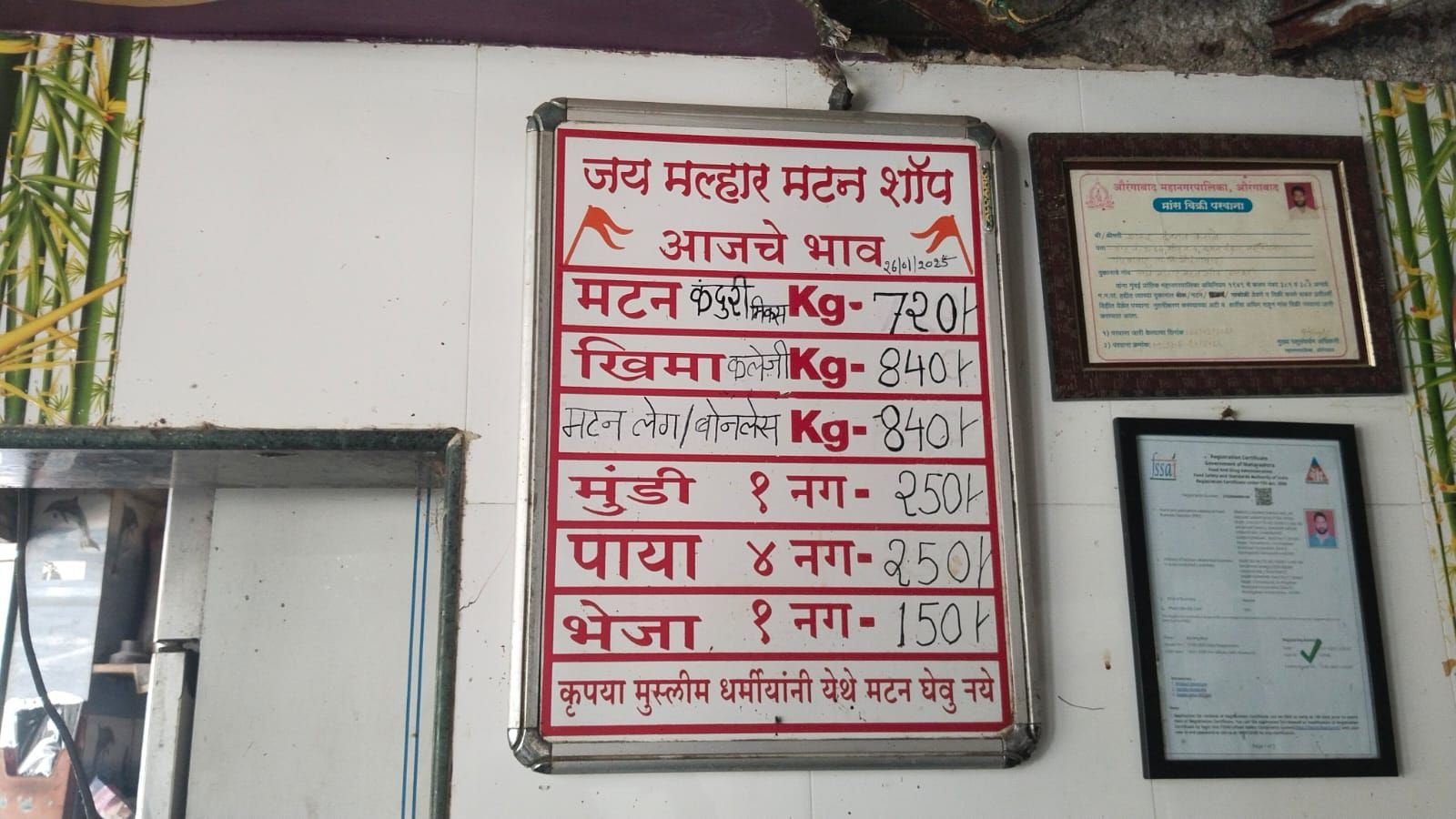 At the bottom of the board, there is a request asking people of the Muslim faith not to buy meat from this shop.  Framed certificates, including an FSSAI registration, are visible on the right. The setting appears to be inside the shop, with tiled walls and a glass display counter.