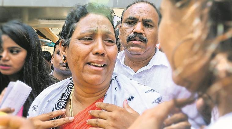 A distressed woman in a red and white saree is seen crying, with her hands on her chest, expressing grief or anguish. Behind her, a man in a white shirt has a serious expression, while several other people, including a woman holding papers, are present in the background. The image captures an intense emotional moment in what appears to be a public or legal setting.