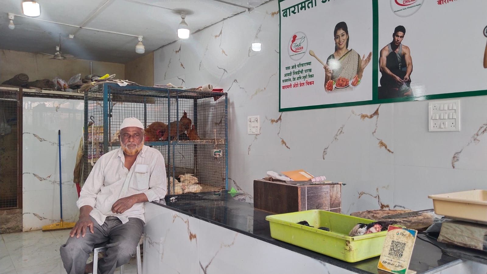 The image shows an elderly man with a white beard and a traditional Muslim skullcap (taqiyah) sitting inside a poultry shop. Behind him, live chickens are kept in metal cages. The shop has clean, white marble-patterned walls and bright lighting. There are posters on the wall promoting "Baramati Agro Chicken," featuring a woman cooking and a fit man, likely emphasizing the quality and health benefits of their chicken. On the counter, there are a few trays, a wooden box, and a QR code for digital payments. In the foreground, a plastic tray contains some freshly cut poultry parts. The overall environment appears organized and well-maintained.